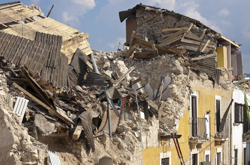 Crumbling buildings with shattered roofs and debris after an earthquake, set against a blue sky. The scene conveys destruction and chaos.